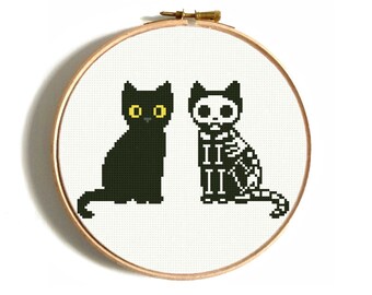 Può includere: Schema di punto croce con due gatti neri, uno con un designo scheletrico. Lo schema è mostrato in un cerchio di ricamo in legno rotondo.
