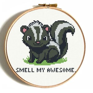 Pode incluir: Bordado em ponto cruz em um bastidor de madeira apresenta um simpático skunk com um padrão de listras pretas e brancas. O skunk está sentado na grama verde, com o texto "SMELL MY AWESOME" abaixo. O bordado está em tecido branco.