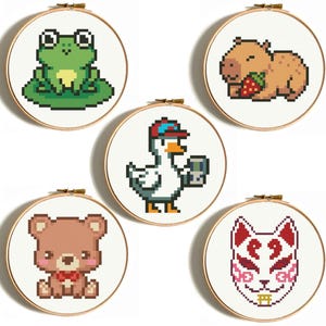 Pode incluir: Cinco bastidores de bordado em pixel art com animais fofos. Um sapo verde, uma capivara com um morango, um pato com boné e dispositivo portátil, um urso marrom com laço vermelho e uma máscara de raposa vermelha e branca.