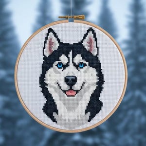 May include: Cross-stitch embroidery of a Siberian Husky dog's face, featuring black and white fur, blue eyes, and a pink tongue. The artwork is displayed in a round wooden hoop, perfect for wall decor. The background is a blurred image of a snowy forest.