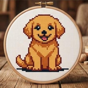 Pode incluir: Um bastidor de bordado circular de madeira contendo um padrão de ponto cruz de um cachorrinho de cor dourada. O cachorrinho é renderizado em um estilo pixelizado com olhos escuros, nariz preto e uma pequena língua rosa sobre tecido branco.