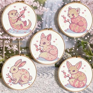 Pode incluir: Cinco bastidores de ponto cruz redondos com animais adoráveis: uma lontra, um panda vermelho, uma raposa, um coelho e um feneco. Cada animal é decorado com padrões de flores de cerejeira e flores, num fundo sereno e desfocado.
