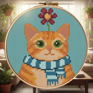 Pode incluir: Bordado em ponto cruz de um gato malhado laranja usando um cachecol azul e branco, com uma flor vermelha brotando de sua cabeça. O gato tem olhos verdes e está sobre um fundo azul claro, emoldurado em um bastidor de madeira.