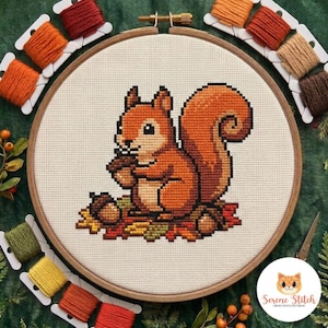 Pode incluir: Um projeto de ponto cruz num bastidor de madeira com um esquilo pixelizado a segurar uma bolota, rodeado por folhas de outono. O desenho é bordado em tecido cor de creme, acompanhado por várias bobines de fio de bordar em tons outonais.
