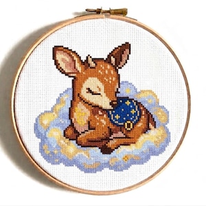 Pode incluir: Um bastidor de bordado circular de madeira contendo um padrão de ponto cruz de um pequeno cervo castanho com manchas brancas, dormindo sobre uma nuvem fofa azul-clara e amarela. O cervo usa uma manta azul-escura decorada com estrelas amarelas.