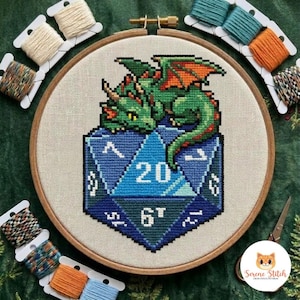 May include: A round wooden embroidery hoop containing a cross-stitch pattern of a green dragon resting on a blue twenty-sided die. The die features white numbers including 20 and 6. Several small spools of colored embroidery thread surround the hoop.