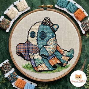 Pode incluir: Um projeto de ponto cruz num bastidor de madeira apresentando uma criatura estilo patchwork com secções estampadas em azul, laranja e branco. A criatura está sentada sobre um pedaço de relva verde. Várias bobinas de fio rodeiam o bastidor.