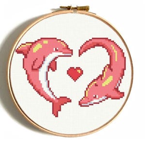 Pode incluir: Um bastidor de bordado em ponto cruz com dois golfinhos cor-de-rosa formando um coração, com um coração vermelho no centro. Os golfinhos têm detalhes amarelos e estão sobre um fundo branco. O bastidor é de madeira com um fecho de latão.