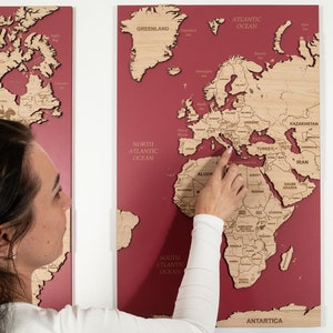 Puede incluir: Un mapa del mundo de madera en tres piezas con fondo rojo. El mapa está dividido en tres secciones, cada una mostrando una parte diferente del mundo. El mapa está montado en una pared y una persona está señalando el mar Mediterráneo.