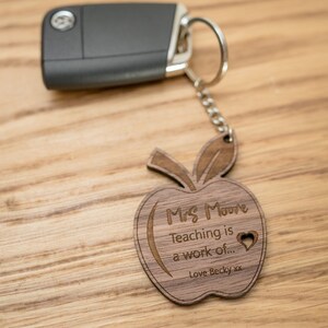 May include: Wooden apple keychain with the text "Mrs. Moore Teaching is a work of..." and a heart. The keychain also includes a silver key ring and a small gray key.