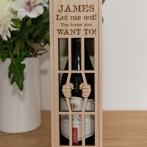 Puede incluir: Una caja de vino de madera con un diseño de celda de prisión. La caja tiene un recorte para una botella de vino y el texto "JAMES Let me out! You know you WANT TO!" está grabado en la parte delantera.