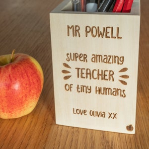 May include: A wooden pencil holder with the text "Mr Powell super amazing teacher of tiny humans Love Olivia xx" engraved on the front.