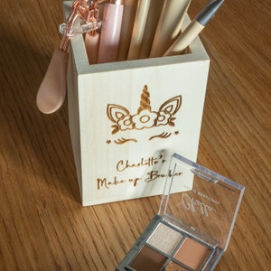 May include: A wooden makeup brush holder with a unicorn design and the text "Charlotte's Make up Brushes". The holder contains makeup brushes, an eyelash curler, and a palette with eyeshadows. The holder is on a wooden surface.
