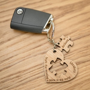 Pode incluir: Um chaveiro de madeira em forma de coração com um recorte de peça de quebra-cabeça. O chaveiro diz "You're the missing piece in my world" e tem os nomes "Elizabeth" e "Anthony" nele.