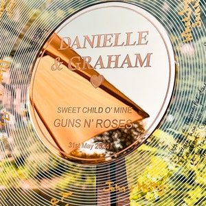 Pode incluir: Uma lembran&ccedil;a de casamento circular em ouro rosa com os nomes "DANIELLE & GRAHAM" gravados. O texto "SWEET CHILD O' MINE GUNS N' ROSES" e a data "31 de maio de 2024" tamb&eacute;m est&atilde;o gravados. O fundo apresenta um design floral.
