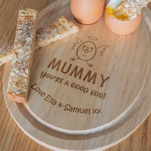 Pode incluir: Bandeja de servir de madeira com um frango de desenho animado bonito e o texto "Mummy you're a good egg! Love Ella & Samuel xx". A bandeja tem duas coqueteleiras de ovo e duas fatias de torradas.