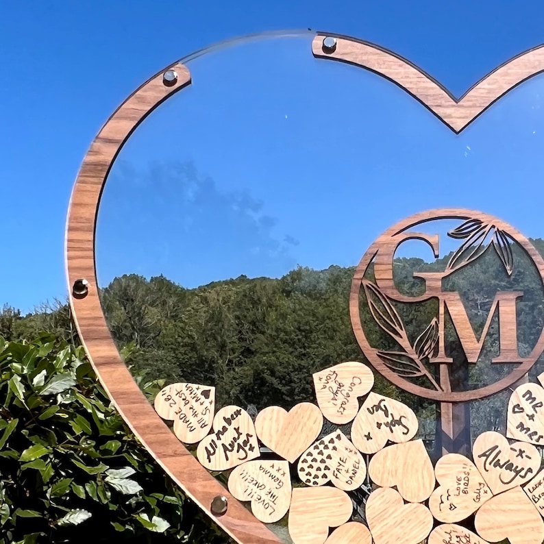 May include: A wooden heart-shaped frame with a clear glass insert. The frame is decorated with a wooden circle with the initials "GM" and leaves. The frame is filled with small wooden hearts with handwritten messages, such as "All the best", "Love", and "Congrats".