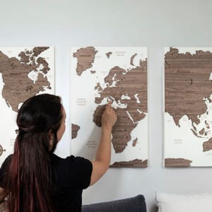 Può includere: Una persona indica una mappa del mondo in legno con sfondo bianco. La mappa è divisa in tre sezioni, con le Americhe, l'Europa e l'Asia su pannelli separati. La mappa è realizzata in legno impiallacciato e ha una finitura in legno naturale.