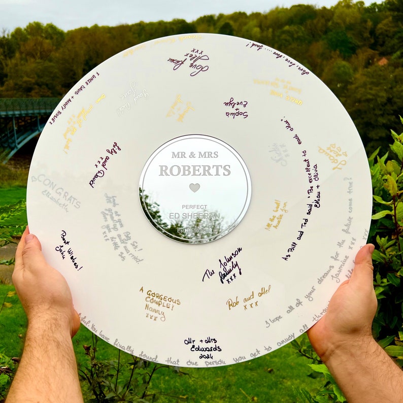 Pode incluir: Um livro de visitas de casamento circular branco com assinaturas em v&aacute;rias cores. O centro apresenta um c&iacute;rculo espelhado com o texto "MR & MRS ROBERTS" e "PERFECT ED SHEERAN". O fundo &eacute; uma cena ao ar livre verde.