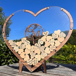 May include: A wooden heart-shaped box with a clear acrylic lid, filled with small wooden hearts. The hearts have handwritten messages on them. The box is on a wooden stand. The box has a wooden plaque with the initials "G" and "M" in a circle.