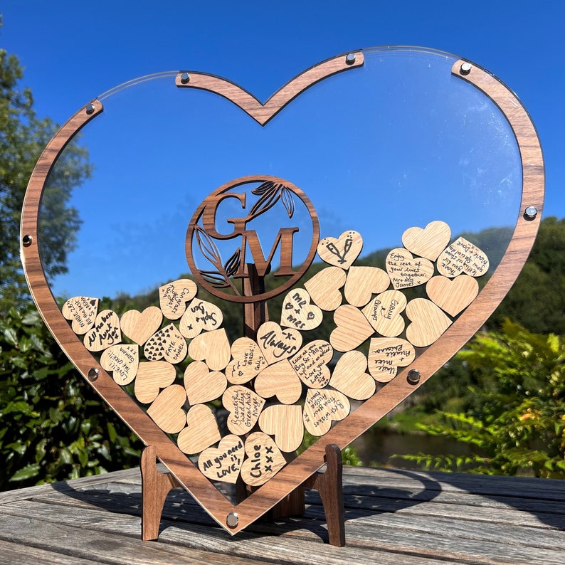 May include: A wooden heart-shaped box with a clear acrylic lid, filled with small wooden hearts. The hearts have handwritten messages on them. The box is on a wooden stand. The box has a wooden plaque with the initials "G" and "M" in a circle.
