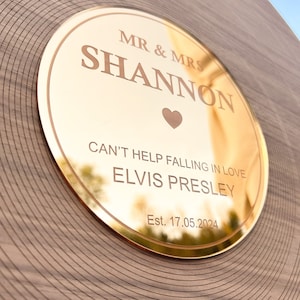 Pode incluir: Placa redonda dourada com os nomes "MR & MRS SHANNON" e a frase "CAN'T HELP FALLING IN LOVE ELVIS PRESLEY". Um s&iacute;mbolo de cora&ccedil;&atilde;o est&aacute; entre os nomes. A data "Est. 17.05.2024" tamb&eacute;m est&aacute; gravada.