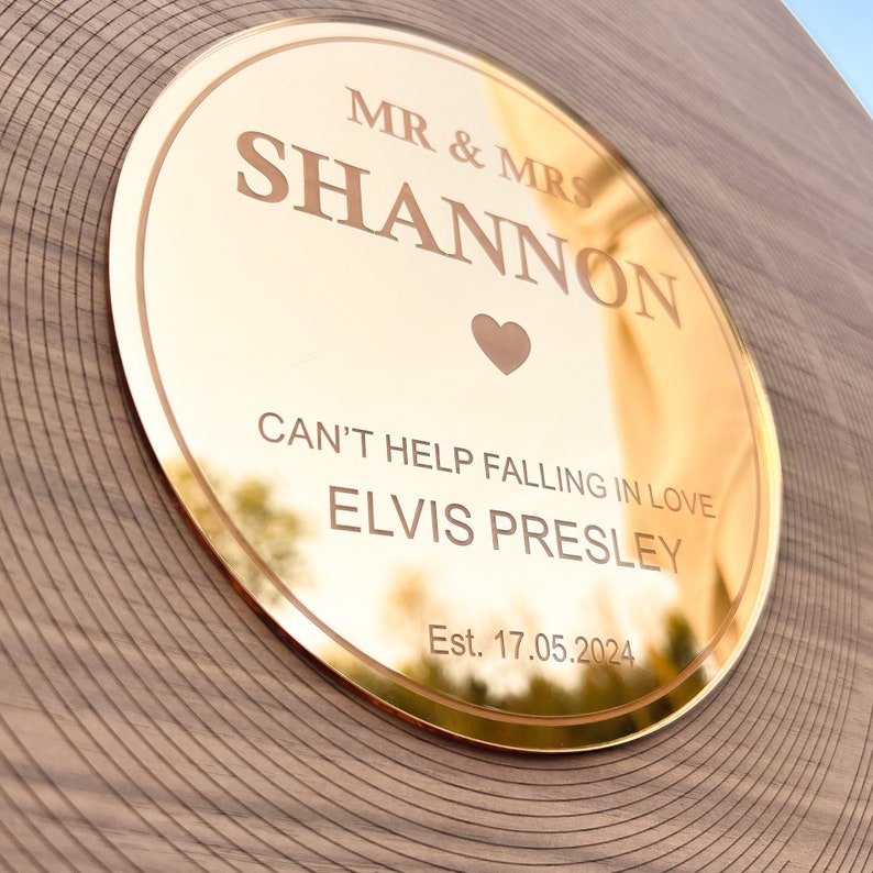 Pode incluir: Placa redonda dourada com os nomes "MR & MRS SHANNON" e a frase "CAN'T HELP FALLING IN LOVE ELVIS PRESLEY". Um s&iacute;mbolo de cora&ccedil;&atilde;o est&aacute; entre os nomes. A data "Est. 17.05.2024" tamb&eacute;m est&aacute; gravada.