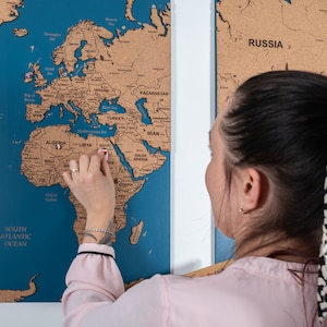以下が含まれることがあります： 青い背景の コルク製の地球儀にピンを刺している女性。地図にはアフリカ、アジア、ヨーロッパの各大陸が表示されています。地図は壁に取り付けられています。