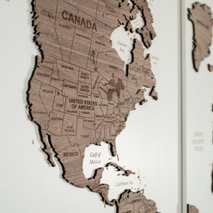 May include: A wooden world map with a raised relief design. The map is divided into continents, with North America highlighted. The map is made of light brown wood and has a white background.