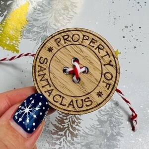 May include: A round wooden button with the text "PROPERTY OF SANTA CLAUS" engraved around the edge. The button has a red and white thread stitched through the center and is attached to a red and white string.