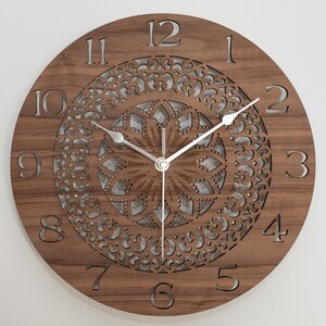 May include: A brown wooden clock with a decorative mandala design and silver hands. The clock face has Roman numerals for the hours and Arabic numerals for the minutes.