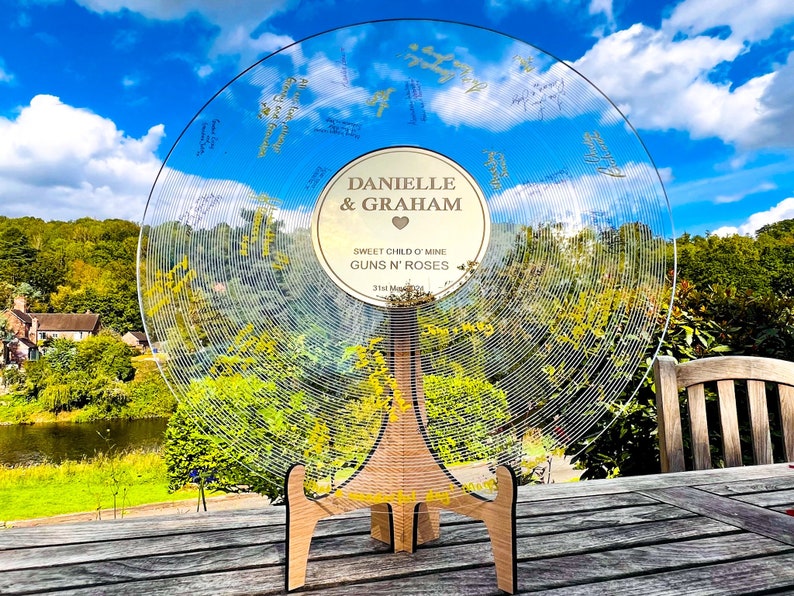 Pode incluir: Um livro de visitas de casamento em acr&iacute;lico transparente em forma de disco de vinil. O disco tem linhas gravadas e &eacute; suportado por um suporte de madeira. O centro do disco tem os nomes "Danielle & Graham" e o t&iacute;tulo da m&uacute;sica "Sweet Child O' Mine".