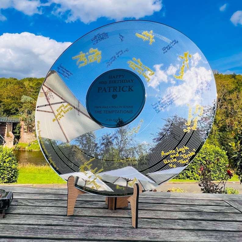 Pode incluir: Um disco de vinil transparente e refletor com um suporte de madeira. O disco tem um centro azul com o texto "HAPPY 60TH BIRTHDAY PATRICK" e assinaturas douradas. O disco reflete o c&eacute;u e a paisagem circundante.