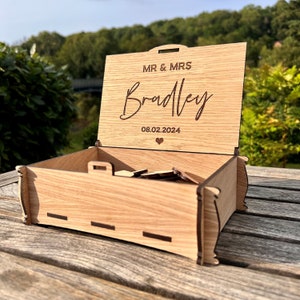 May include: A wooden box with a hinged lid, personalized with the text "MR & MRS Bradley 08.02.2024" and a small heart symbol. The box is open, revealing a small stack of wooden pieces inside.