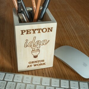 Op de afbeelding: Een houten potloodhouder met de naam Peyton gegraveerd op de voorkant. De houder heeft een gloeilampgrafiek en de tekst "idea" en "Genius at Work".