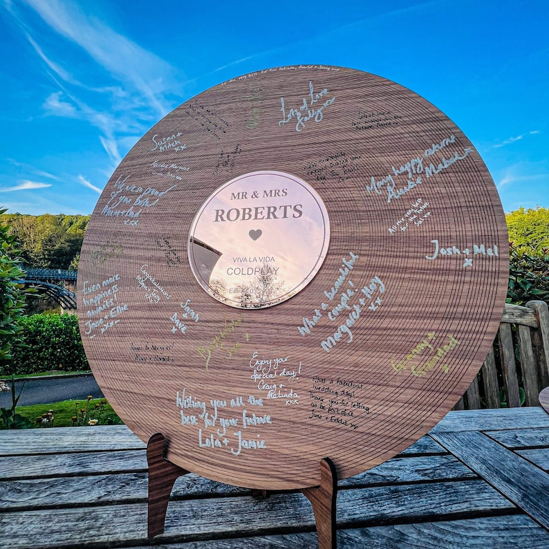 Pode incluir: Uma placa de madeira grande e redonda com um centro espelhado, gravada com "MR & MRS ROBERTS" e "VIVA LA VIDA COLDPLAY". A placa &eacute; coberta por mensagens e assinaturas manuscritas e &eacute; suportada por um suporte de madeira.