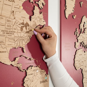 May include: A red wooden world map with push pins marking locations. The map is divided into three sections, with North America, South America, and Europe visible. A hand is placing a push pin on the map.