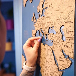 Peut inclure: Une carte du monde en bois avec les contours et les noms des pays, montée sur un fond bleu. Une personne place une pièce de la carte dans son espace correspondant. La carte est un objet décoratif.