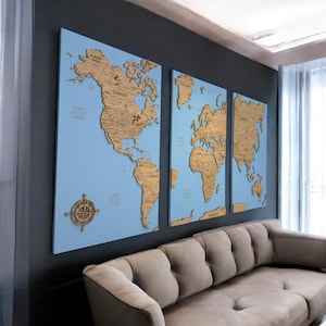 May include: A three-piece wooden world map wall art set with a light blue background. The map is divided into three sections: North America, Europe, and Asia. Each section is raised and textured, giving the map a 3D effect. A compass is included in the bottom left corner of the North America section.