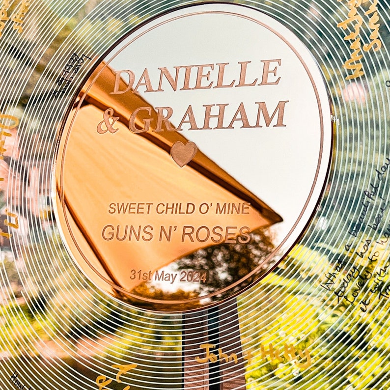 Pode incluir: Uma lembran&ccedil;a de casamento circular em ouro rosa com os nomes "DANIELLE & GRAHAM" gravados. O texto "SWEET CHILD O' MINE GUNS N' ROSES" e a data "31 de maio de 2024" tamb&eacute;m est&atilde;o gravados. O fundo apresenta um design floral.