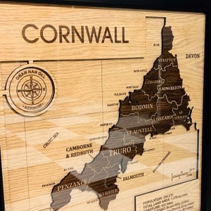 Può includere: Una mappa in legno della Cornovaglia, Inghilterra, con il nome della contea inciso in alto. La mappa è incorniciata in nero e presenta una rosa dei venti con la frase in lingua cornica "Onan Hag Oll" che significa "Uno e Tutti".