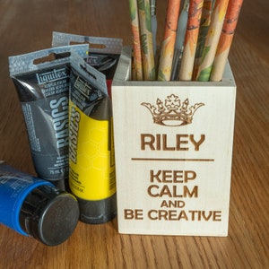 Könnte beinhalten: Ein hölzerner Stiftehalter mit dem Namen "Riley" und dem Text "Keep Calm and Be Creative", der auf der Vorderseite eingraviert ist. Der Halter ist mit verschiedenen Buntstiften und Farbtuben gefüllt.