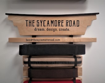 May include: A wooden wall-mounted belt rack with the text "THE SYCAMORE ROAD dream. design. create." and the email address "ben@thesycamoreroad.com". The rack has five belts hanging on it, including a black belt, a tan belt with a black stripe, a brown belt, and a red belt.