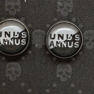 May include: A pair of black and white round button pins with the text "UNUS ANNUS" printed on them. The pins are on a black background.