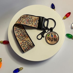 May include: A beige and black wristlet keychain with a black clasp and a round pendant. The wristlet features a repeating alphabet and light design. The pendant has the word "RUN" with a string of lights. The keychain is on a white surface with colorful light bulbs.