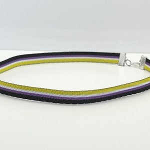 Op de afbeelding: Een choker-ketting met non-binaire pride. De choker heeft een zwart, paars, wit en geel gestreept patroon. Het heeft een zilverkleurige sluiting en ketting voor verstelbare maat.