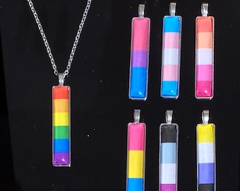 May include: A silver chain necklace with a rectangular pendant featuring the rainbow pride flag. There are also seven other pendants, each featuring a different pride flag, including the transgender, bisexual, pansexual, and non-binary flags.