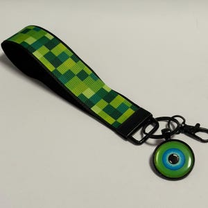 May include: A green pixelated wrist lanyard with a black clasp and a round pendant. The pendant features a green border and a blue and black eye design. The lanyard is approximately 8 inches long.