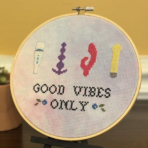 May include: A cross-stitch embroidery hoop with the words "GOOD VIBES ONLY" in black thread. The design includes four colorful symbols and is set against a tie-dye background. The hoop is displayed on a small black easel.
