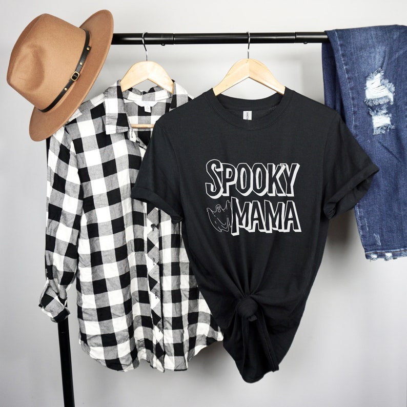 May include: Black t-shirt with white text that reads "Spooky Mama" and a ghost graphic. The shirt is on a hanger with a plaid shirt and blue jeans in the background.